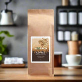 A brown paper bag of Excalix mushroom coffee is displayed on a wooden table in a cozy kitchen setting. The label on the bag features the Excalix logo and reads "Excalix Coffee, Mushroom Coffee Fusion." The background includes blurred kitchen elements such as jars and a coffee pot, creating a warm and inviting atmosphere.