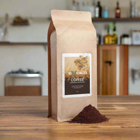 A brown paper bag of Excalix mushroom coffee is displayed on a wooden table in a cozy kitchen setting. The label on the bag features the Excalix logo and reads "Excalix Coffee, Mushroom Coffee Fusion." The background includes blurred kitchen elements such as jars and a coffee pot, creating a warm and inviting atmosphere.